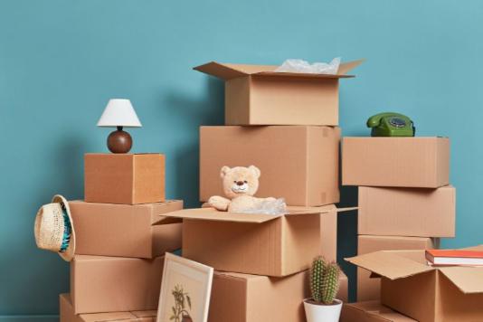 Cardboard boxes piled up with household items displayed on top, a lamp, a hat, a picture, a phone a teddy, a plant